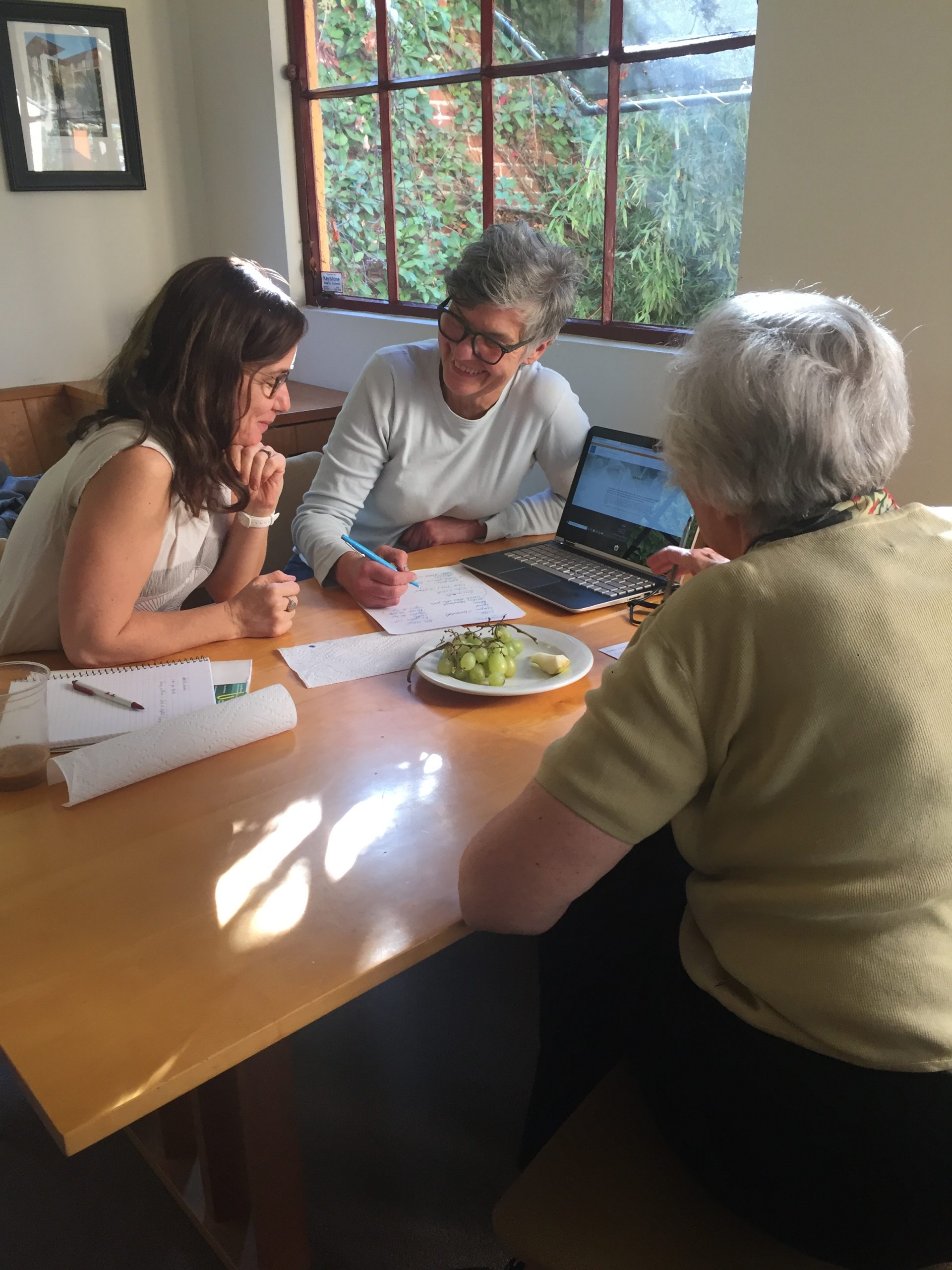 Three women seated around a table with a laptop, smiling and talking.